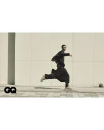 East Asian man in a black jacket and flowing trousers running past a cream paneled wall with a GQ logo in the corner.