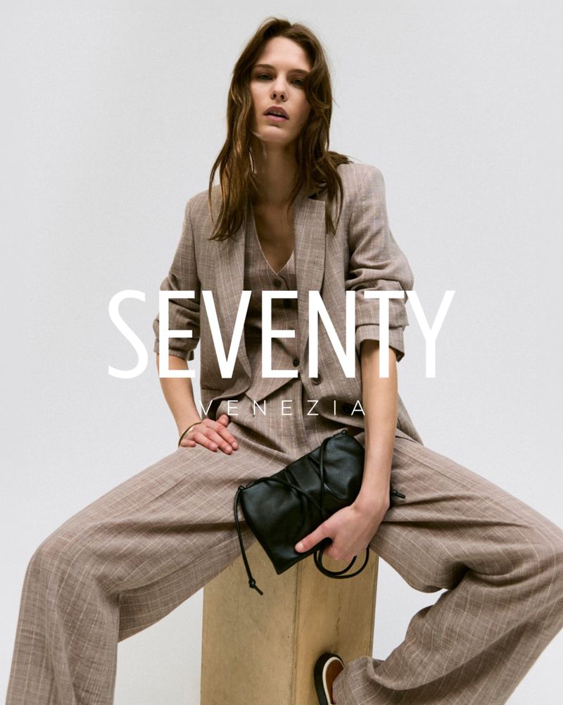 Fashion editorial shot of a brunette woman in a brown pantsuit sitting on a wooden block and holding a black bag against a white backdrop.