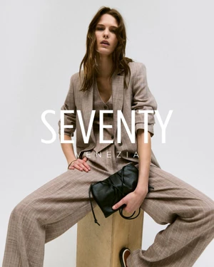 Model Coline Leclere seated wearing a taupe three-piece tailored suit and holding a black leather clutch against a white background.
