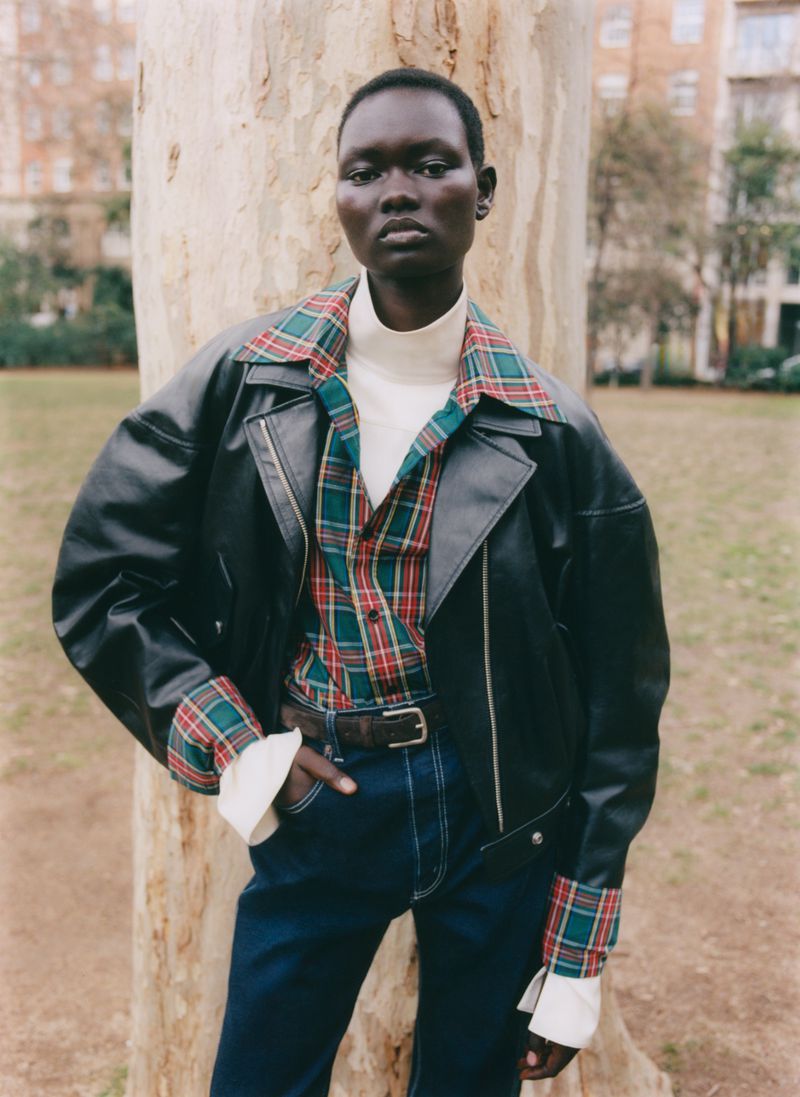 Model Ateta Jok wearing an oversized black leather jacket layered over a plaid shirt and white turtleneck in front of a tree trunk.
