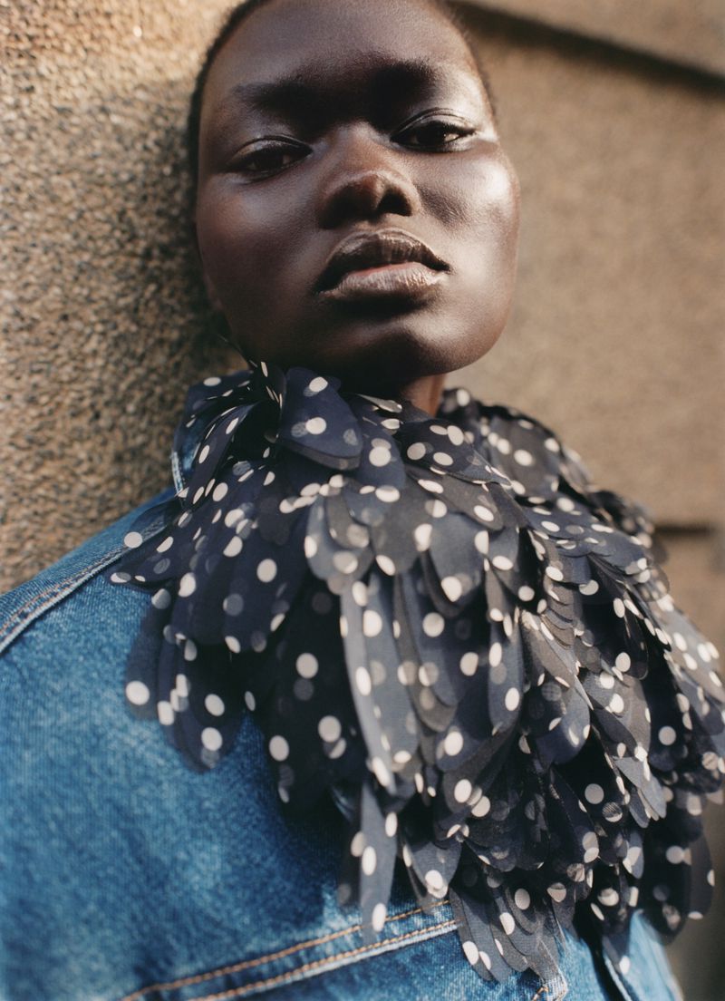 Close-up portrait of model Ateta Jok wearing a sheer polka-dot ruffled collar and blue denim jacket against a textured wall.