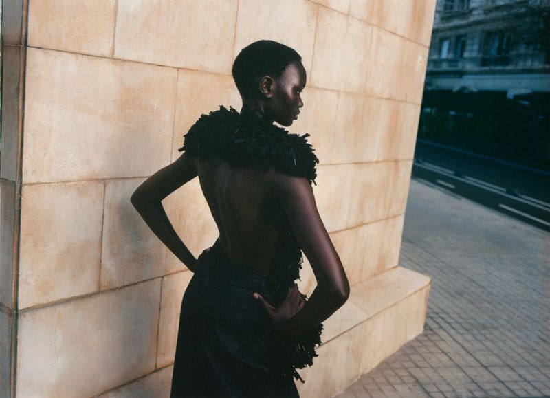 Model Ateta Jok poses outdoors in a black backless textured fringe piece against a warm stone architectural wall.
