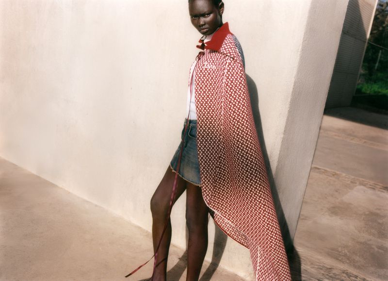 Model wearing a patterned red and white cape and denim miniskirt standing against a pale architectural wall outdoors.