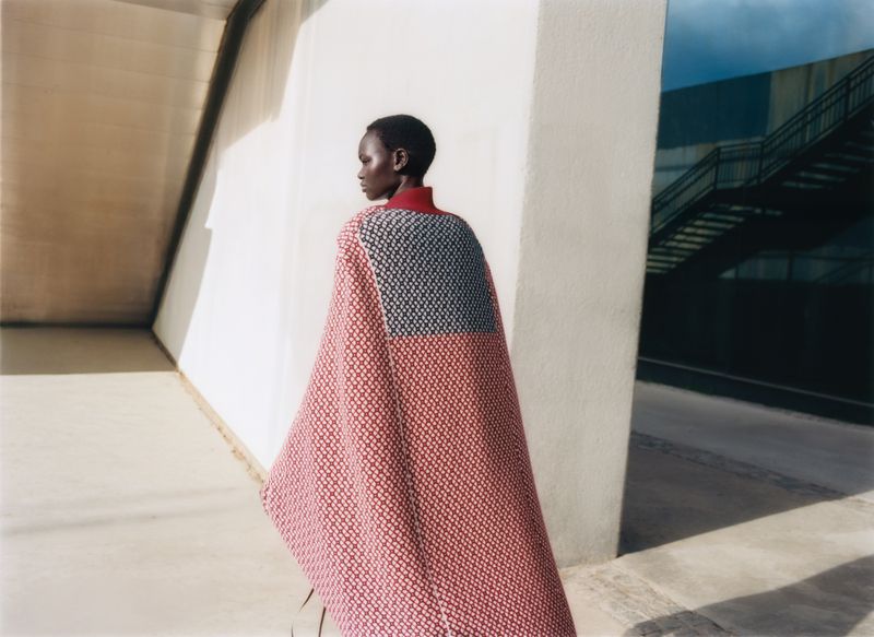 Model wearing patterned red cape standing outdoors near concrete architecture in Vogue Polska editorial
