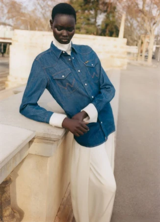 Ateta Jok leans against a stone balustrade wearing a blue denim shirt over a white turtleneck and cream trousers.
