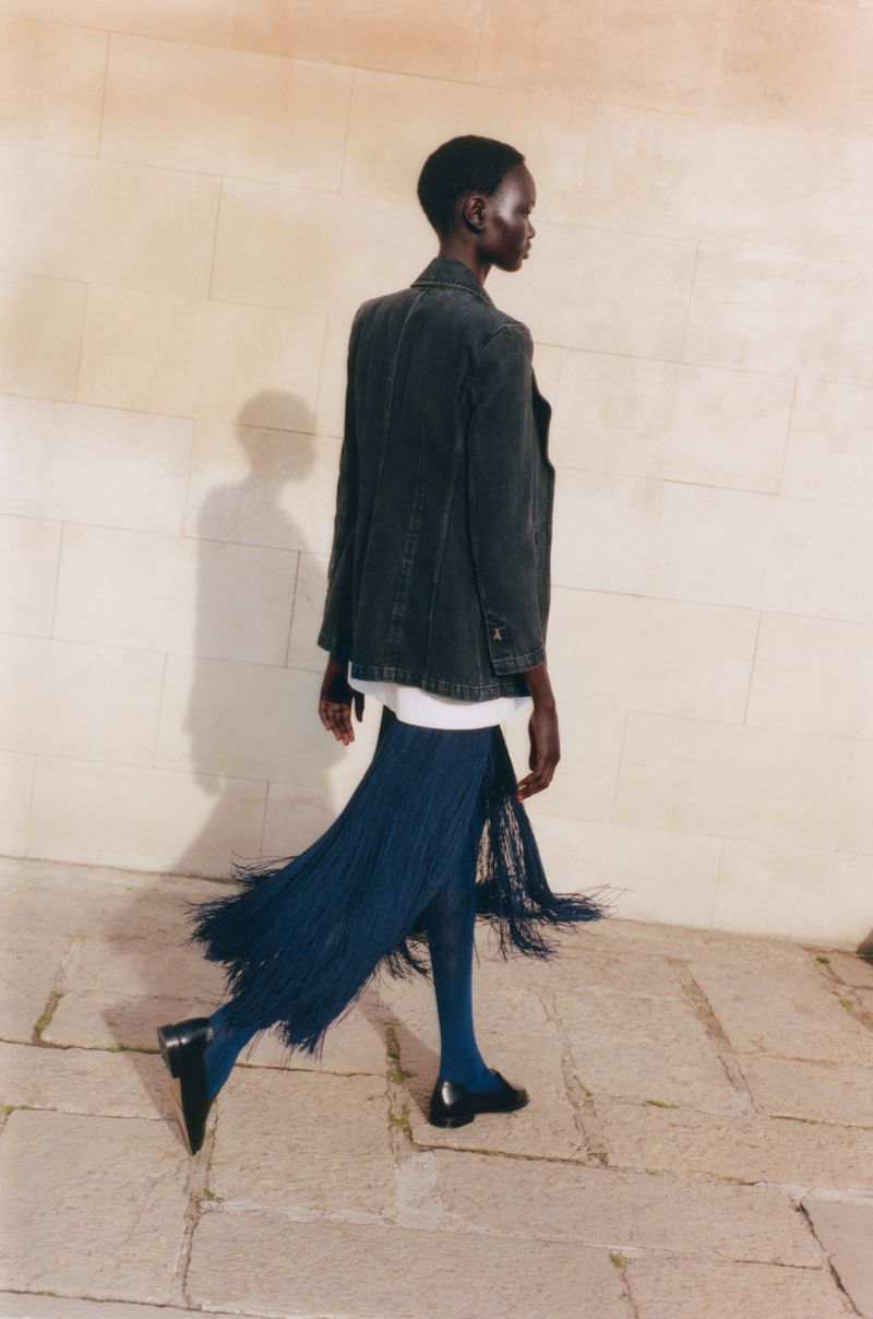 Model Ateta Jok walks past a stone wall wearing a dark blazer, navy fringe skirt, blue tights, and black loafers in natural sunlight.