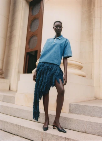 Model Ateta Jok wearing a short-sleeve denim polo shirt and an asymmetric deep blue fringe skirt on stone architectural steps.