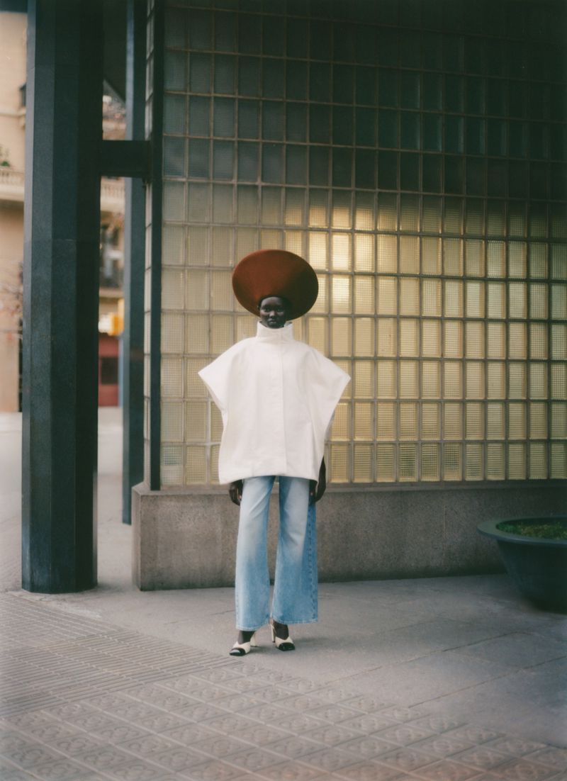 Model wearing a wide white top, blue jeans, and a large round hat standing before a glass brick wall.