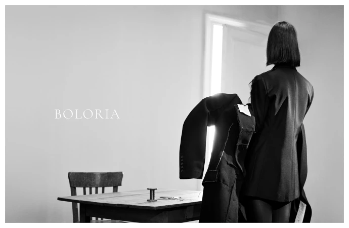 Monochrome image of a woman from behind holding a tailored black coat near a wooden table inside a sparse room.