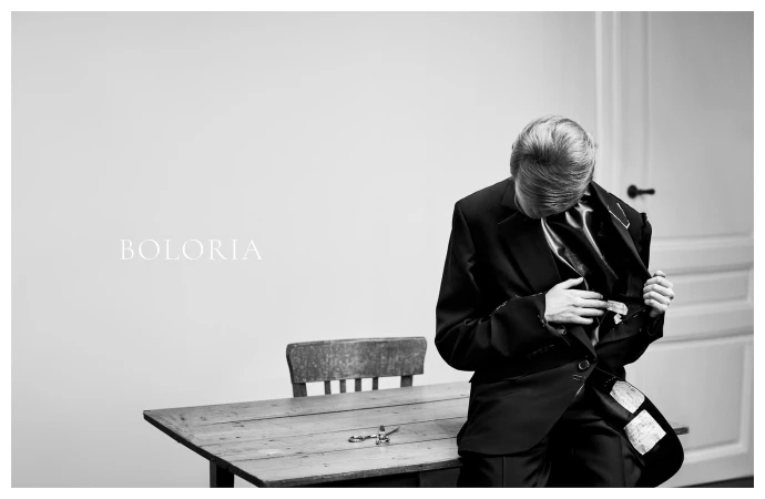 Person in a dark suit seated on a wooden table adjusting a jacket lapel in an interior room