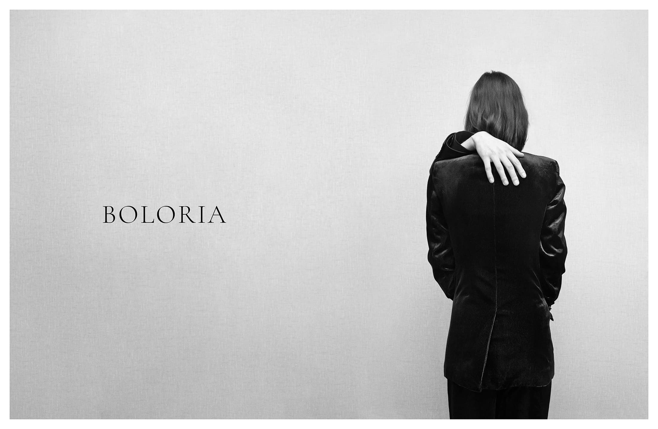 Monochrome fashion ad with a model facing away wearing a dark velvet jacket with a hand over their shoulder beside Boloria branding.