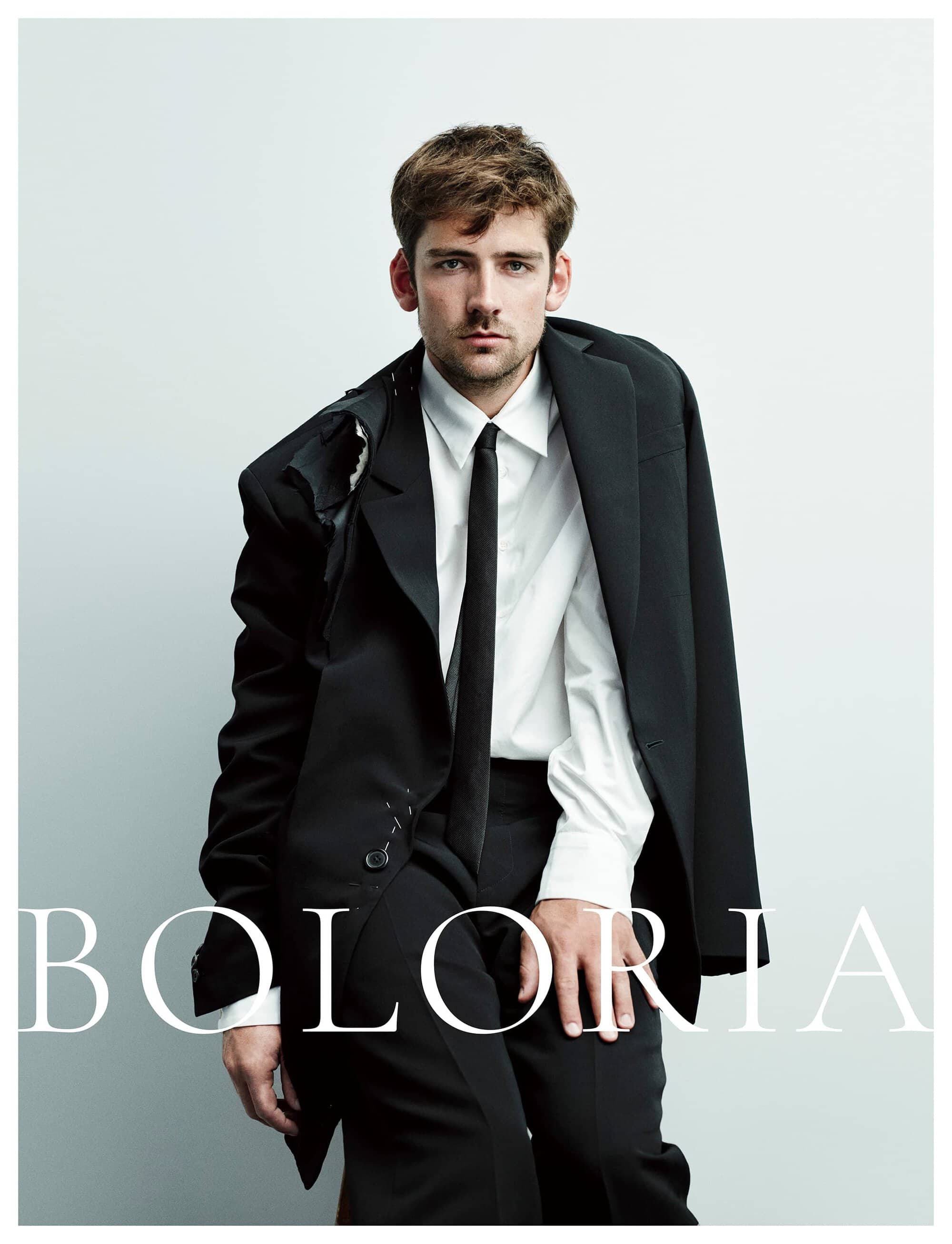 Man wearing a black suit jacket, white button-down shirt, and black tie against a seamless white background with Boloria text