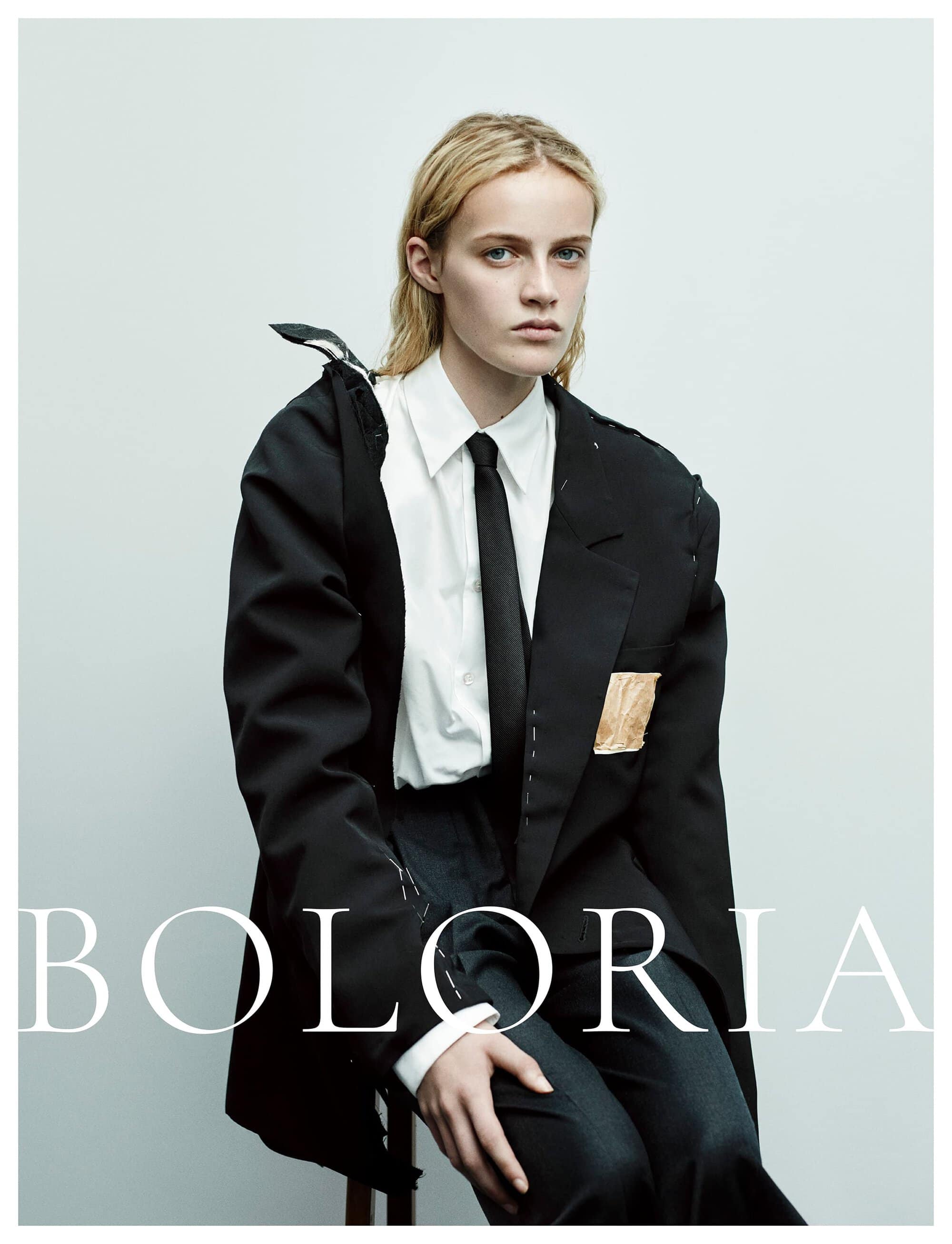 Model wearing a deconstructed black blazer, crisp white shirt, and black tie seated against a pale studio background for Boloria.