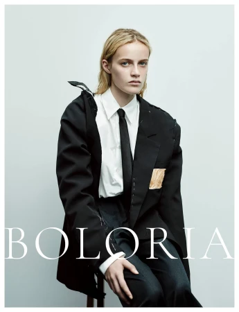 Model wearing a deconstructed black blazer, crisp white shirt, and black tie seated against a pale studio background for Boloria.