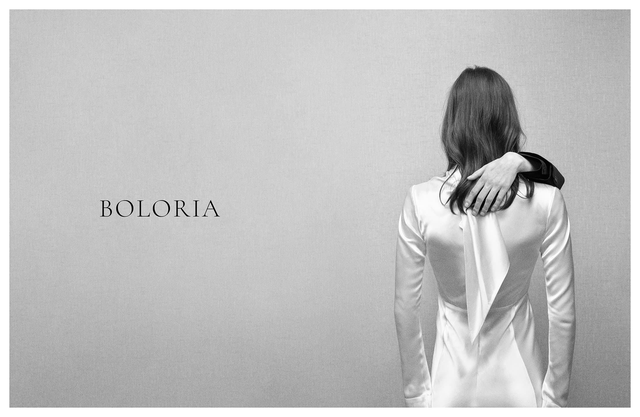 Model seen from behind wearing a white satin dress against a textured light grey backdrop with Boloria logo