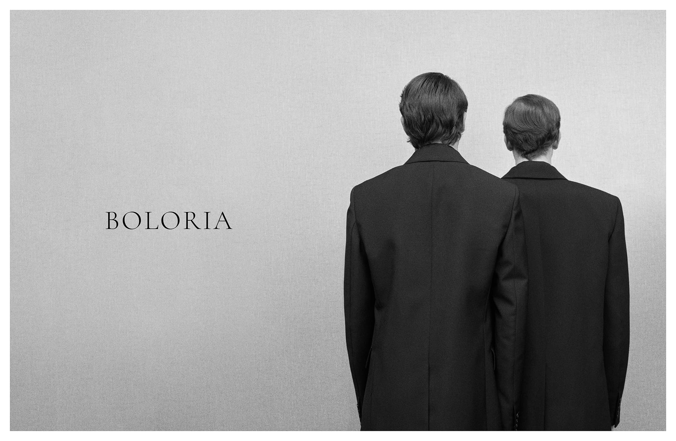 Two models wearing tailored black blazers standing with their backs to the camera against a grey textured studio backdrop with Boloria text.