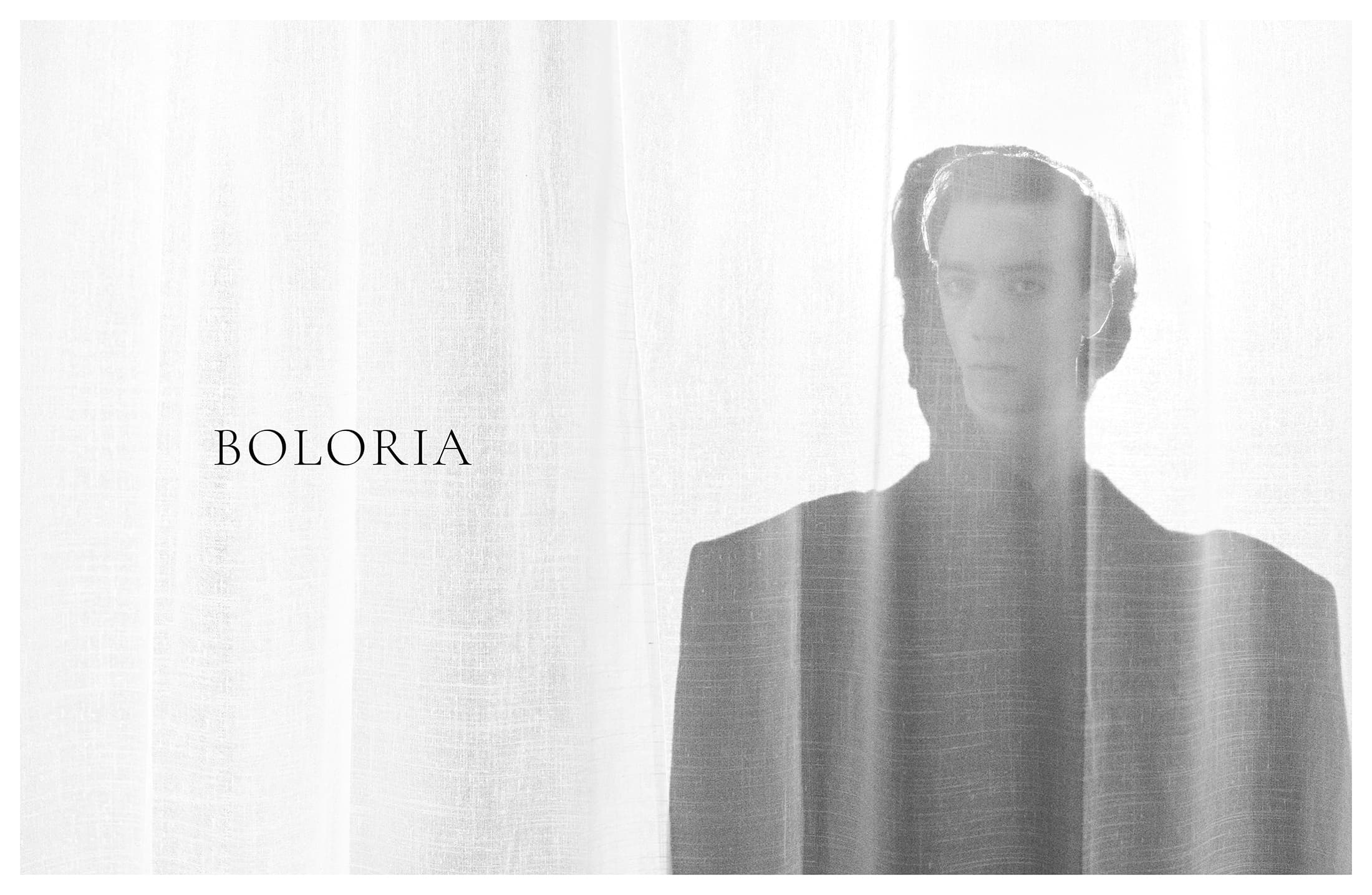Monochrome portrait of a person in a suit, partially obscured by a translucent white curtain. Soft light creates a serene mood.