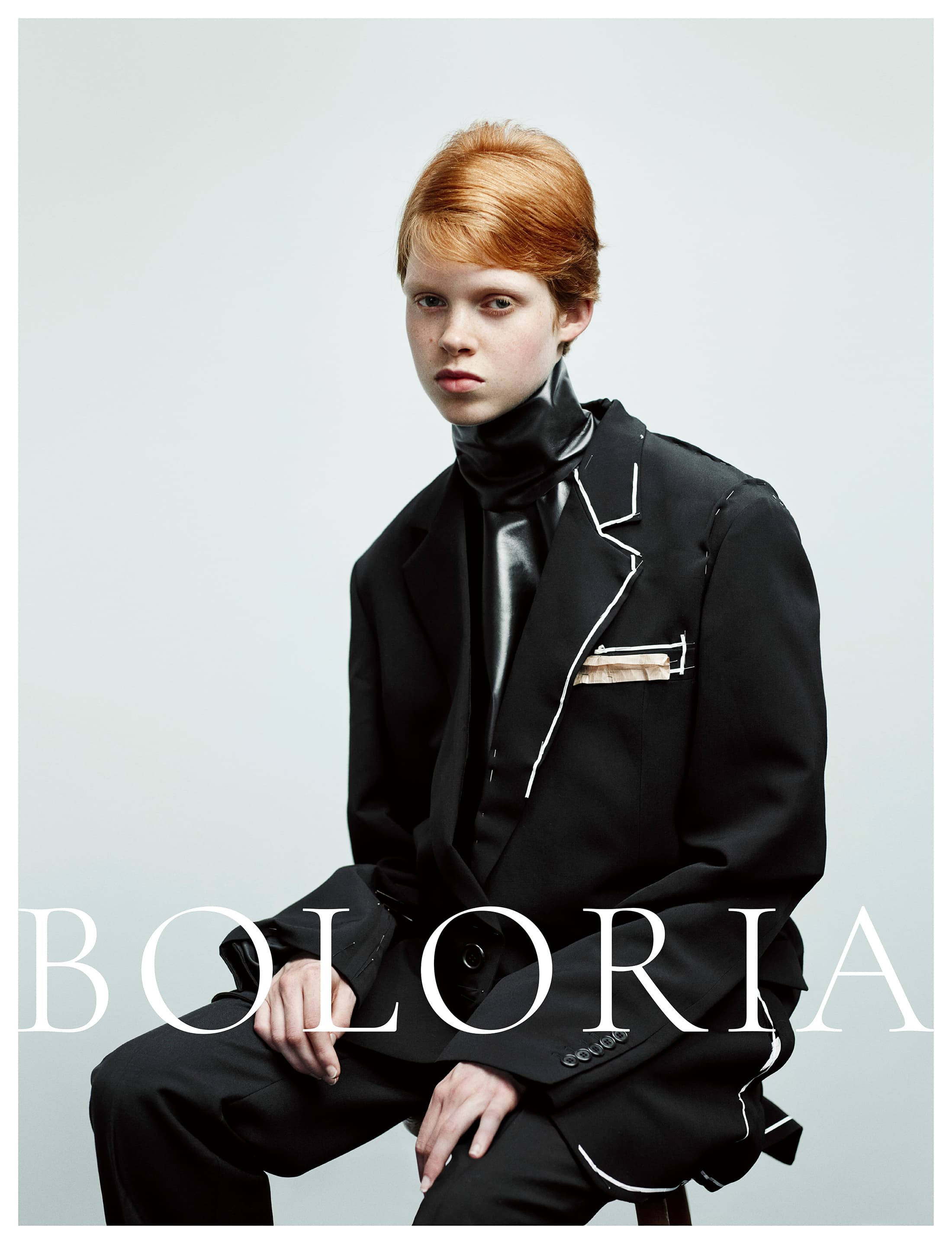 Model with short red hair wearing an oversized black suit with white piping details seated against a pale grey studio background.
