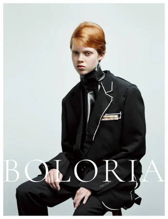 Model with short red hair wearing an oversized black suit with white piping details seated against a pale grey studio background.