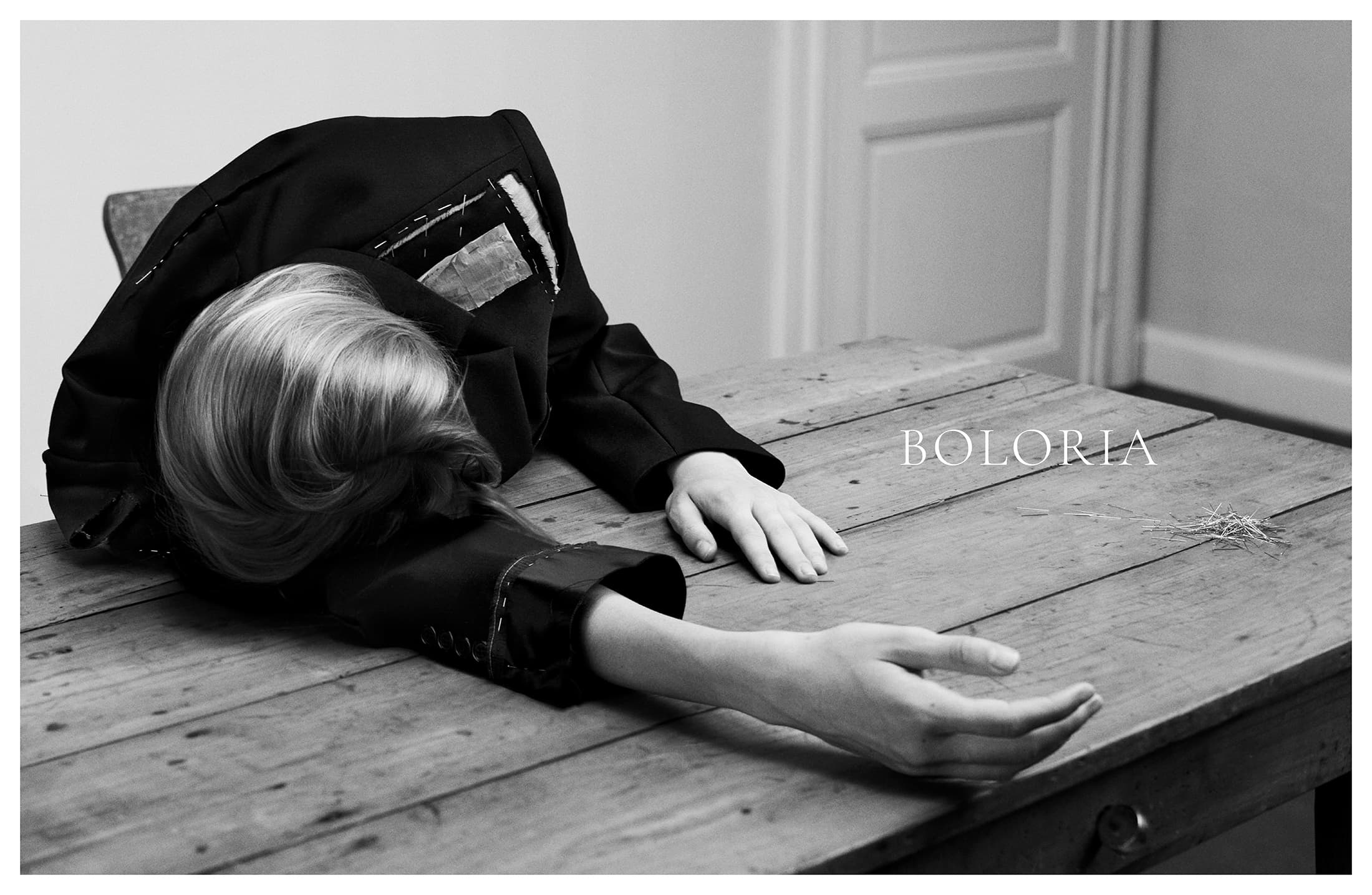 Monochrome image of a model slumped face down across a wooden table wearing a deconstructed black jacket beside the Boloria logo.