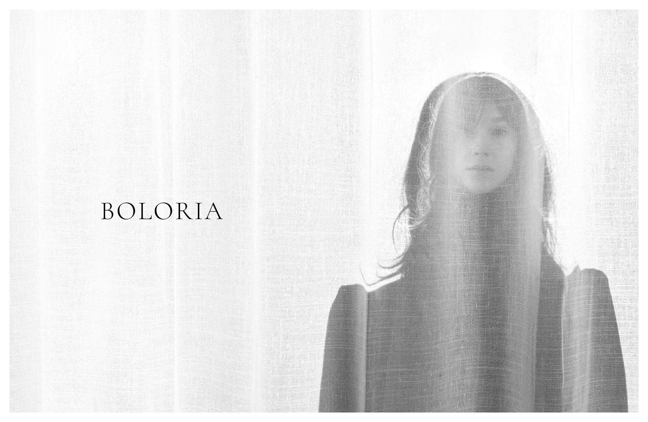 Model standing behind a sheer white curtain wearing a dark garment with the brand name Boloria visible on the left side