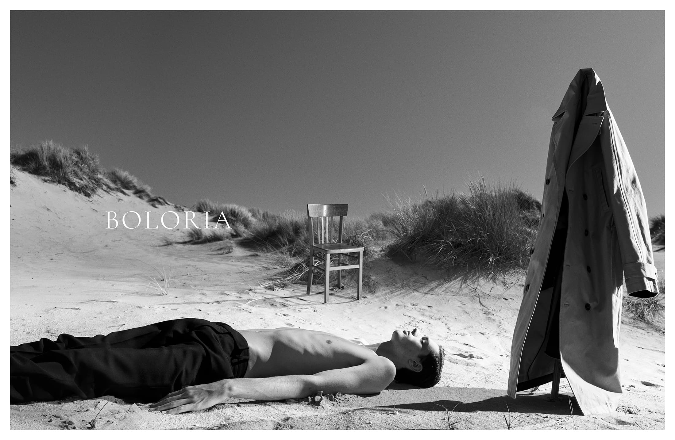 Monochrome fashion campaign image featuring a man lying on a sandy beach next to an empty chair and a standing trench coat