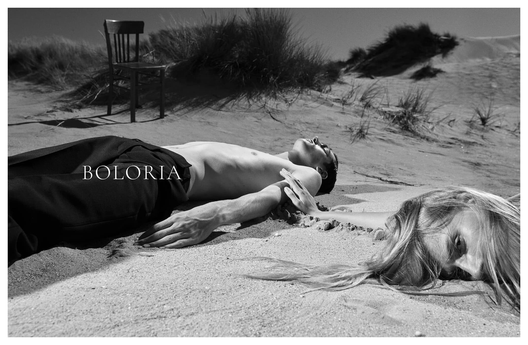 Monochrome fashion campaign image featuring models lying on sandy dunes with a wooden chair in the background