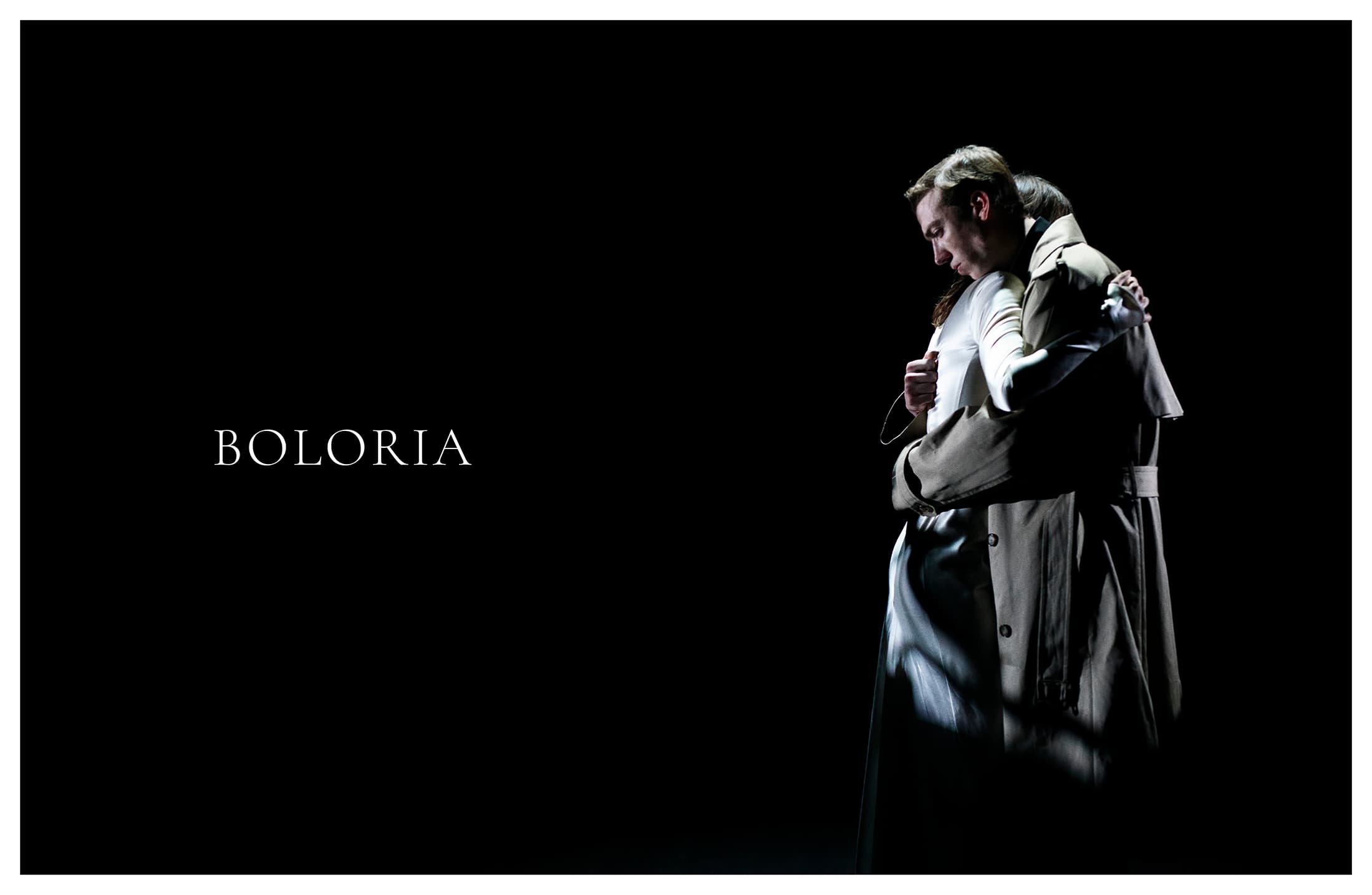 A man in a tan trench coat embraces a person in a white satin gown against a dark background beside the white Boloria logo.