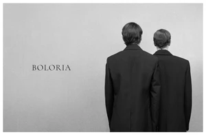 Two models wearing tailored black blazers standing with their backs to the camera against a grey textured studio backdrop with Boloria text.