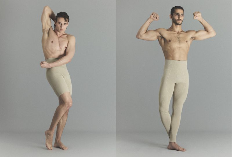 Two shirtless male models posing in high-waisted beige knit shorts and leggings against a neutral grey studio backdrop.