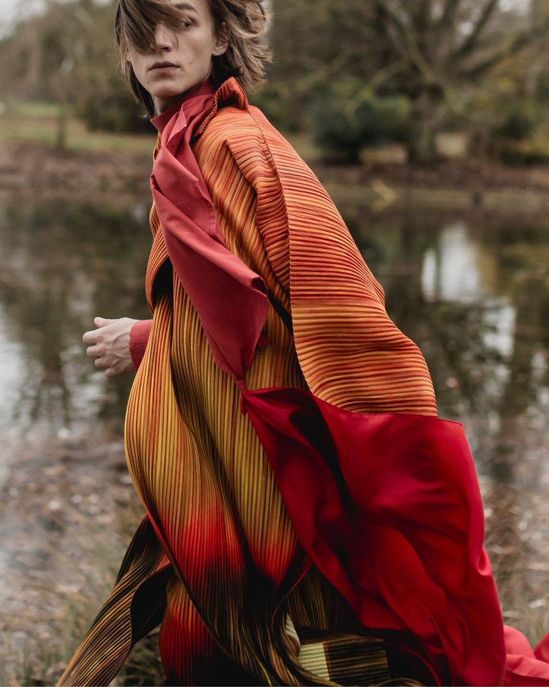 Model in red and orange pleated coat walking outdoors near a body of water for The Greatest Magazine editorial