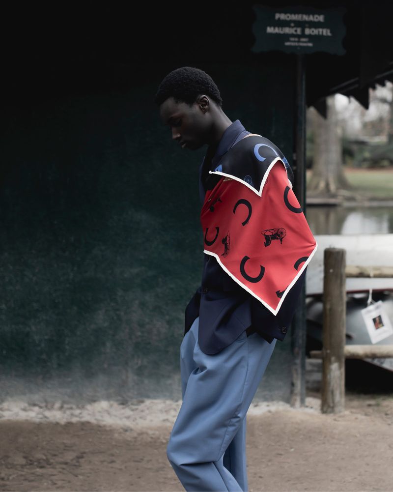 Model Bakary Cisse in a navy jacket, patterned red scarf, and blue trousers standing outdoors by a park promenade sign.