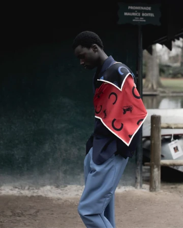Model Bakary Cisse in a navy jacket, patterned red scarf, and blue trousers standing outdoors by a park promenade sign.