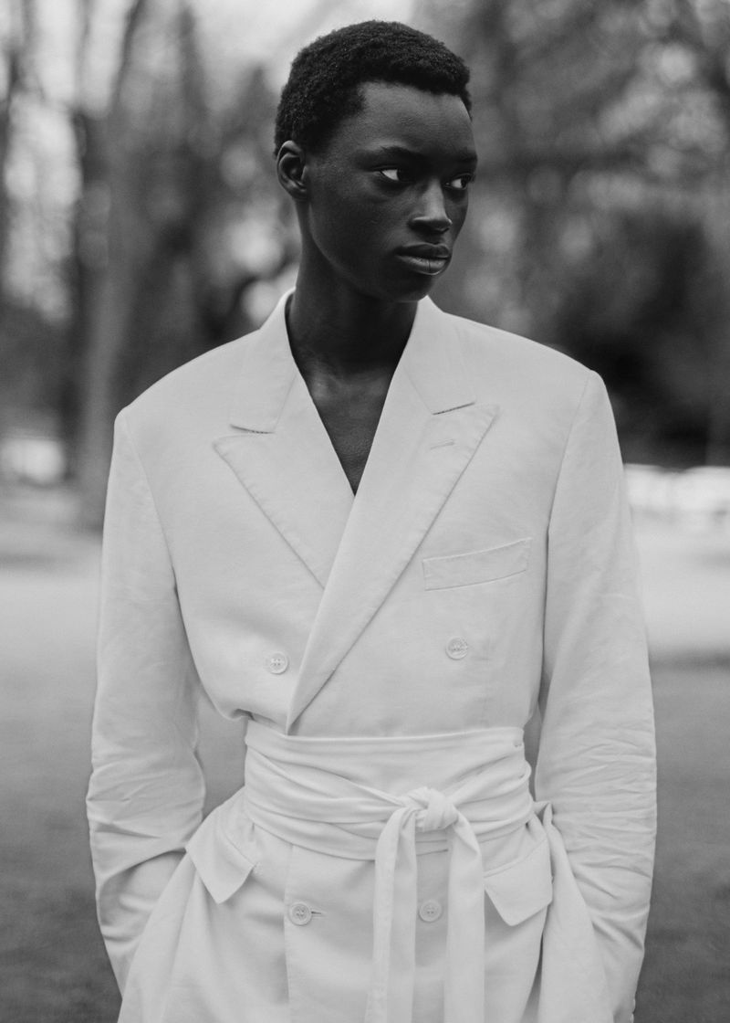 Black-and-white fashion portrait of Bakary Cisse wearing a belted white double-breasted jacket outdoors.