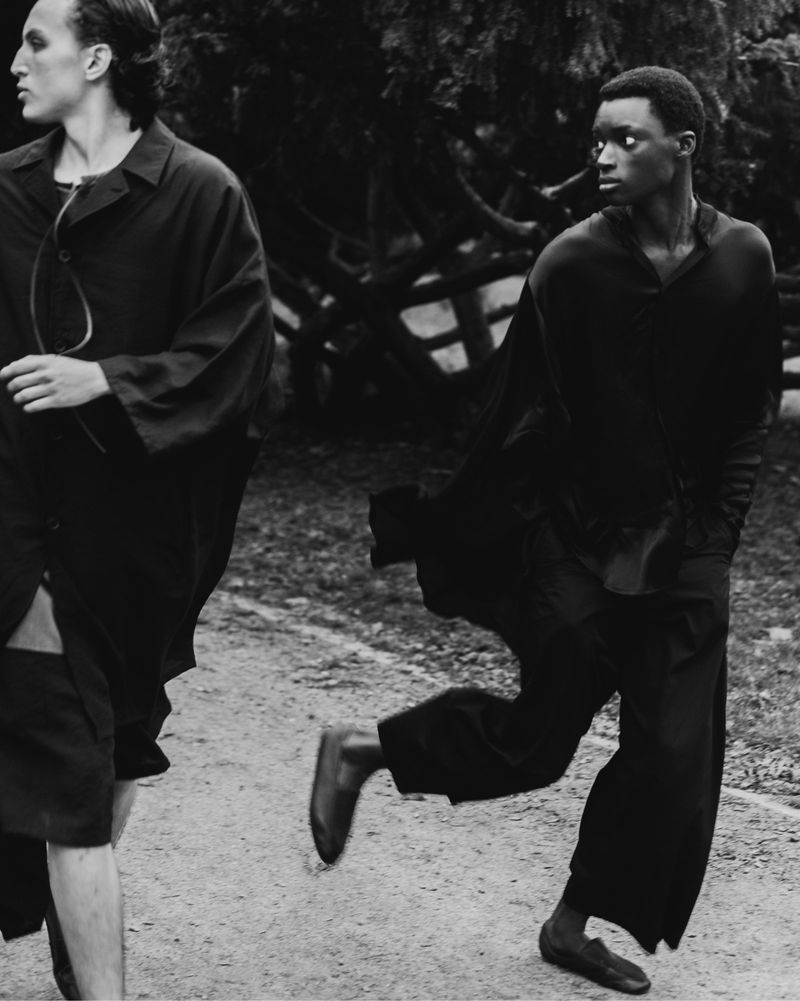 Monochrome fashion shot of two adult males in dark clothing outdoors; one male running in mid-stride on a dirt path surrounded by trees.