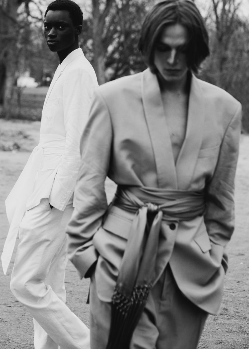 Black and white photo of models Michael Zielinski in a belted blazer and Bakary Cisse in a white suit outdoors in a park.