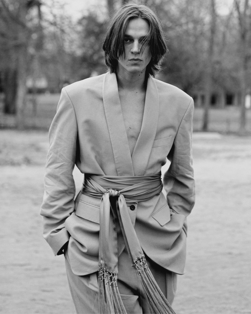 Black and white portrait of a male model wearing an oversized blazer with a wide tied sash standing outdoors