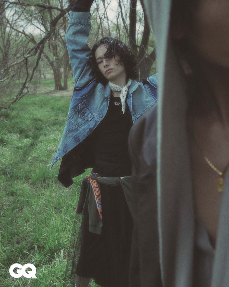 Stylish young man in denim jacket poses outdoors with a hooded figure in the foreground, set against a backdrop of trees and greenery.