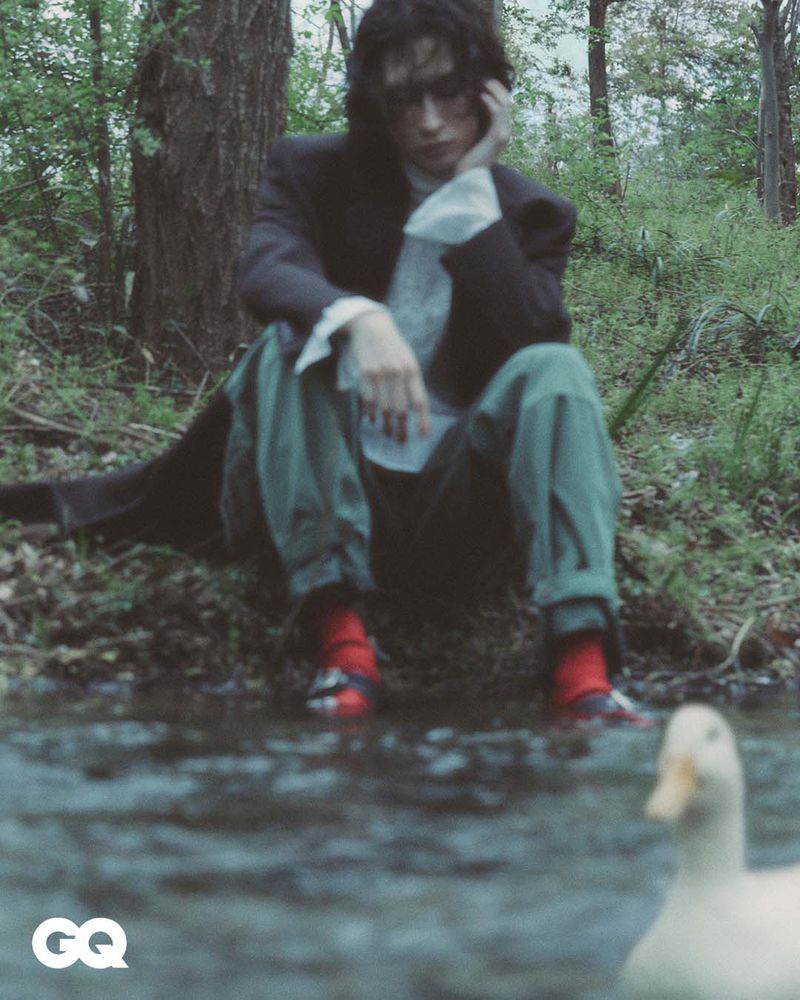 Dark-haired person sits in forest wearing a black coat and red socks; a duck swims in the stream in the foreground, GQ logo is visible.