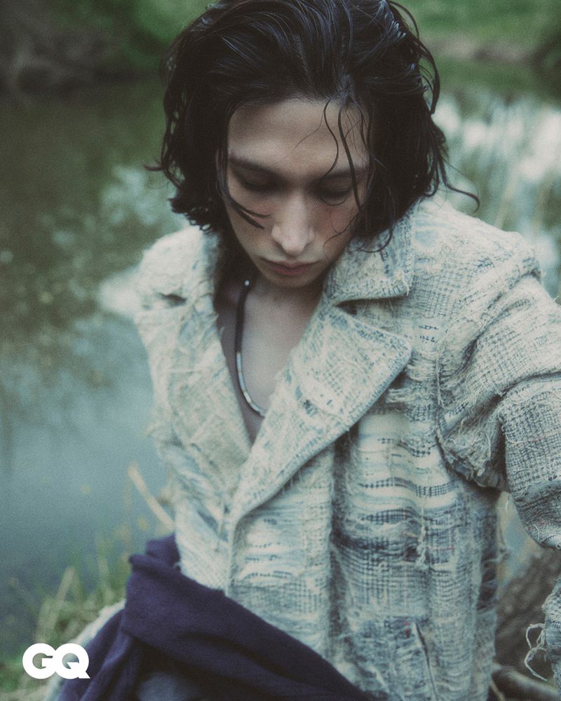 Outdoor fashion portrait of an adult male wearing a textured jacket near a body of water, captured in soft, diffused natural lighting.