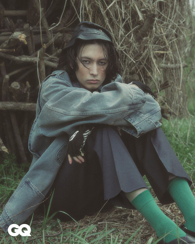 A young adult with a serious expression sits in the grass near a brush pile, wearing a denim jacket and dark pants, styled for a magazine.