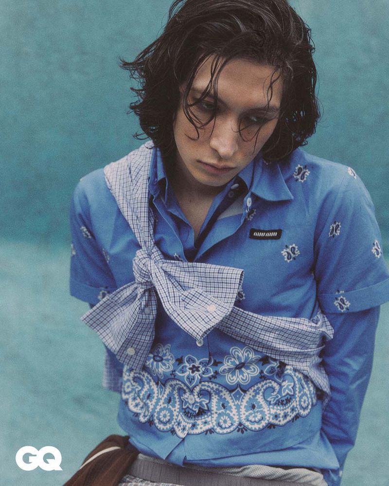Portrait of a young adult with long dark hair wearing a patterned blue shirt and white checkered scarf, set against a blue backdrop.