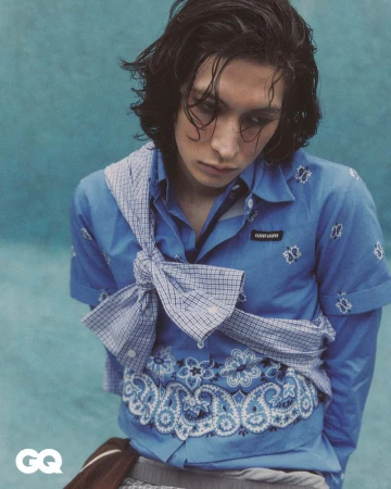 Portrait of a young adult with long dark hair wearing a patterned blue shirt and white checkered scarf, set against a blue backdrop.