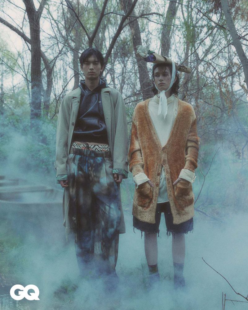 Two models in fashionable outerwear pose outdoors in a surreal forest setting with fog. A fashion editorial shot for GQ magazine.