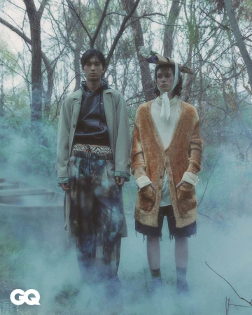 Two models in fashionable outerwear pose outdoors in a surreal forest setting with fog. A fashion editorial shot for GQ magazine.