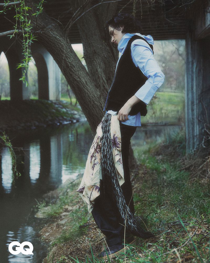 Stylish man in outdoor fashion shoot, wearing a light blue shirt and black vest, posing on a riverbank with concrete bridge behind him.