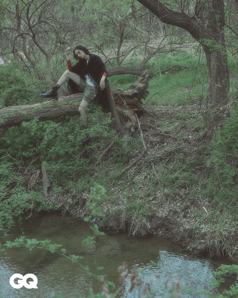 A young adult poses on a fallen tree trunk in a green forest alongside a stream. The natural setting creates a moody atmosphere.