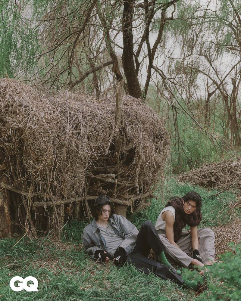 Two adult men pose outdoors near a makeshift structure of twigs and branches, wearing earth-toned casual clothing in a natural setting.