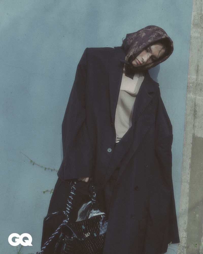 Fashionable adult leans against wall in oversized coat with chain-strap handbag, wearing patterned head covering and light top.