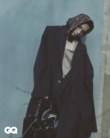 Fashionable adult leans against wall in oversized coat with chain-strap handbag, wearing patterned head covering and light top.